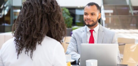 Friendly diverse business colleagues working on project over cup of coffee. Business man sitting at table opposite of his female colleague, listening to her and smiling. Business lunch concept