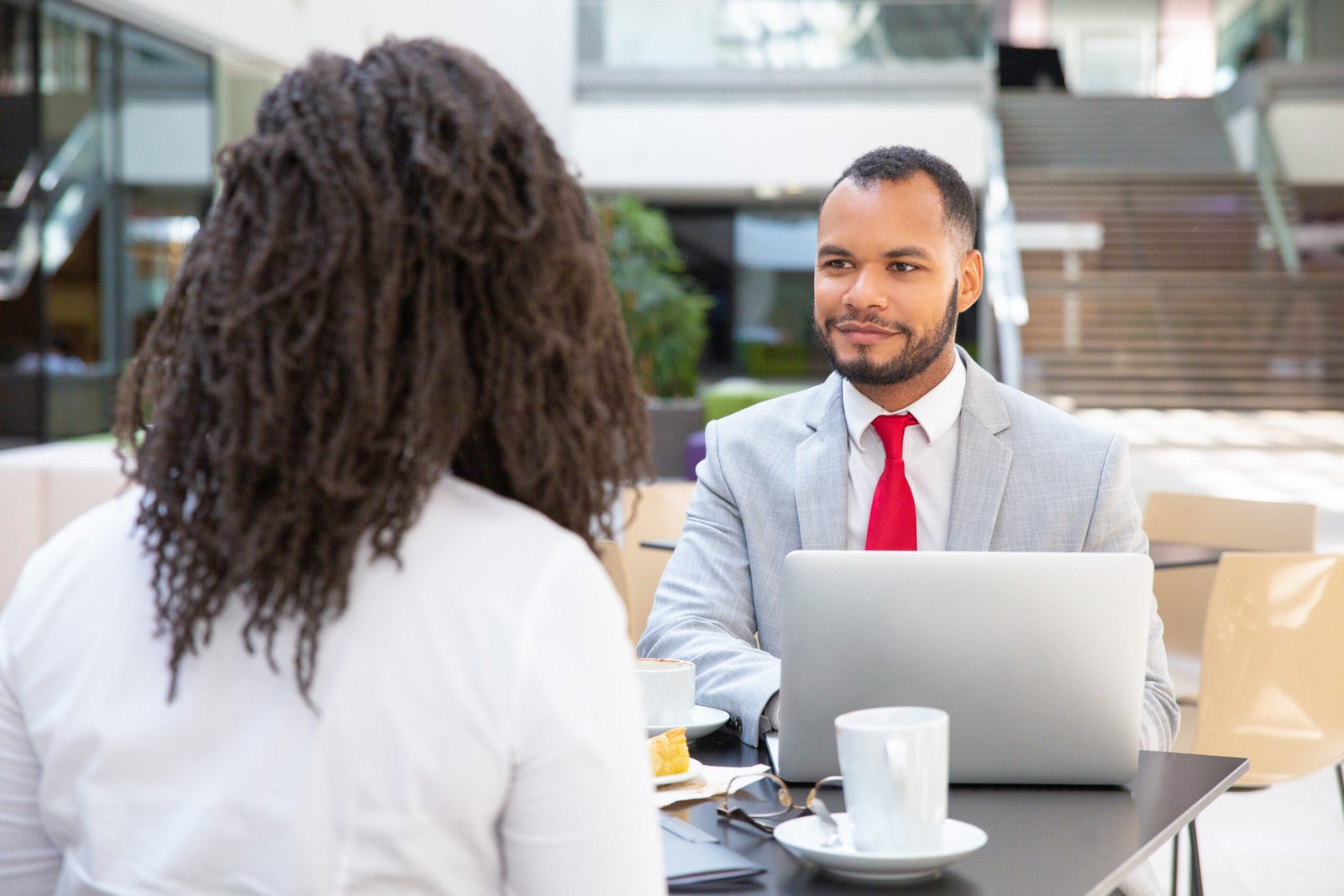 Friendly diverse business colleagues working on project over cup of coffee. Business man sitting at table opposite of his female colleague, listening to her and smiling. Business lunch concept